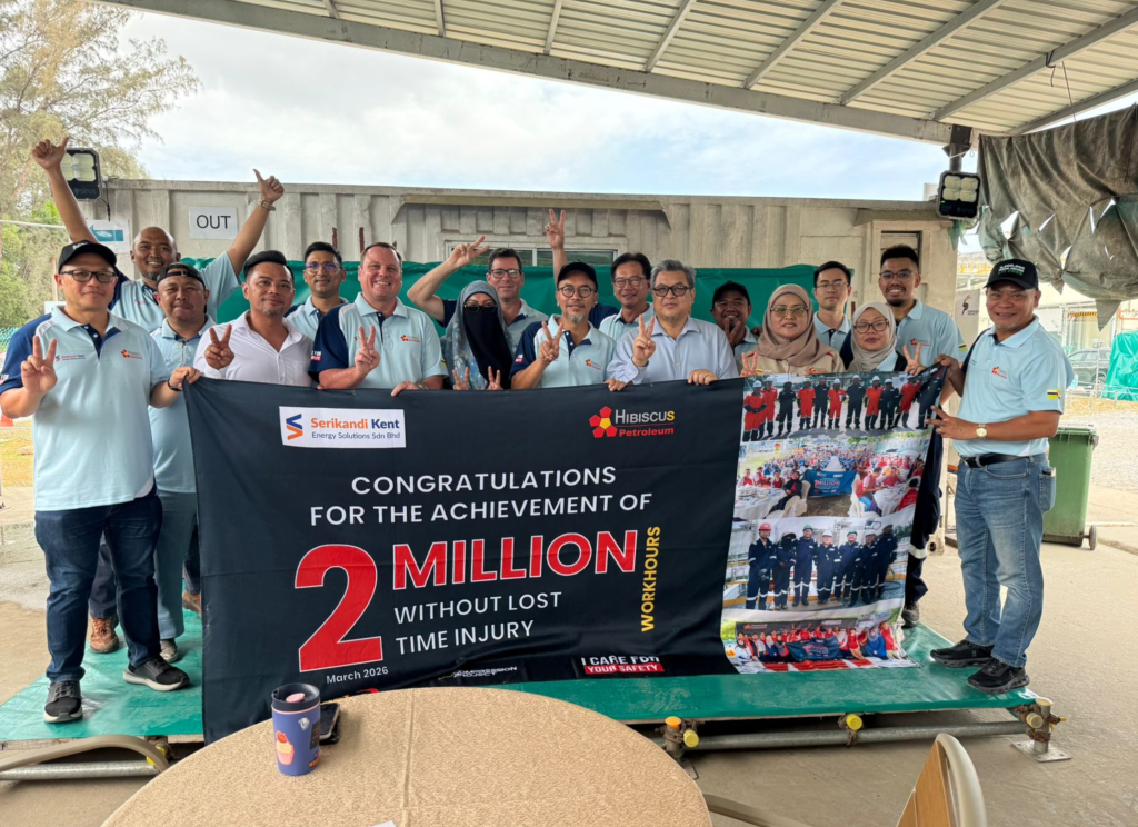 A group of 16 employees and contractors stand together under a covered outdoor worksite area, smiling and posing with peace signs and thumbs-up gestures while holding a large congratulatory banner. The banner, branded with Serikandi Kent Energy Solutions and Hibiscus Petroleum logos, celebrates the achievement of “2 Million Work Hours Without Lost Time Injury” in March 2026. Several team members wear matching light blue company polo shirts, while others wear casual work attire. A photo-collage panel highlighting team and safety milestones is displayed on the right side of the banner.